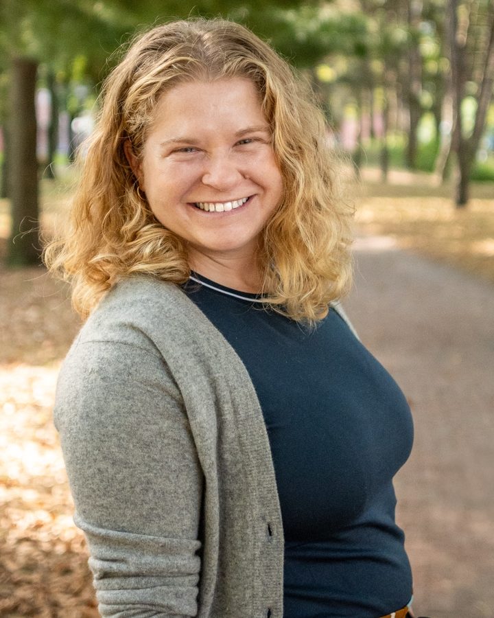 Aly Silver smiling on a tree-lined path, wearing a gray cardigan
